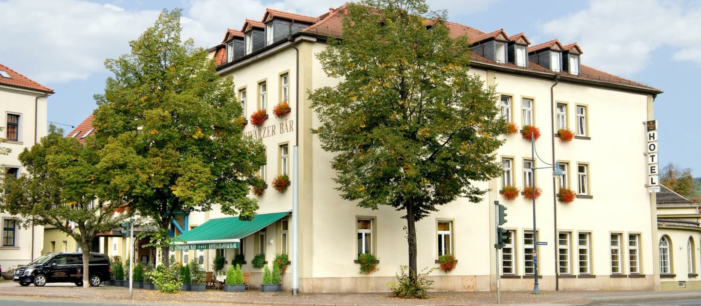 Dreistöckiges Hotelgebäude mit rotem Ziegeldach, mehreren Fenstern mit Blumenkästen und grünem Markisenüberdach am Eingang, umgeben von Bäumen an einer Straßenkreuzung in Jena.