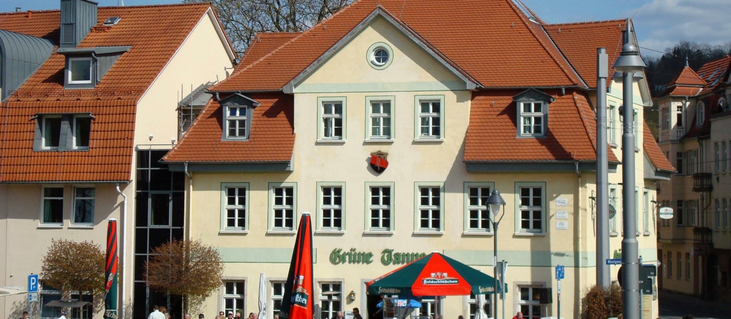 Dreigeschossiges Gebäude mit rotem Satteldach und Schriftzug »Grüne Tanne« an der Fassade, davor Terrasse mit Sonnenschirmen und zahlreichen Menschen, im Hintergrund weitere Gebäude und blauer Himmel mit Wolken