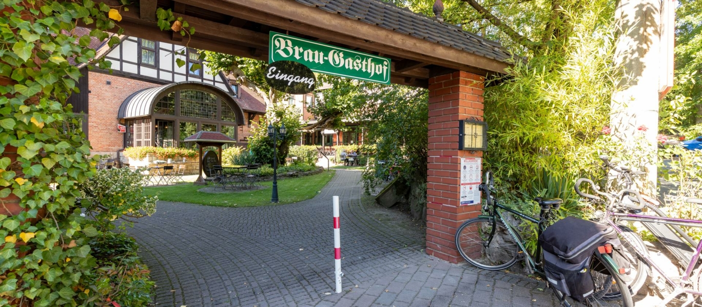 Eingang zu dem Brau-Gasthof Papiermühle mit grünem Schild über einem Torbogen aus roten Ziegeln, davor ein gepflasterter Weg und mehrere Fahrräder an einem Fahrradständer.