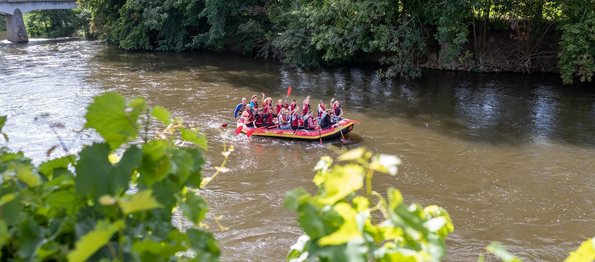 Gruppe von Menschen in einem Schlauchboot auf der Saale umgeben von Bäumen und einer Brücke im Hintergrund