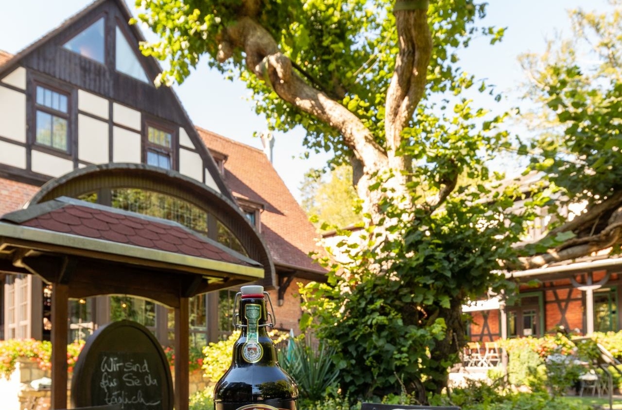Bierflasche und zwei Biergläser mit Schaum stehen auf einem Holztisch im Außenbereich des Braugasthofs Papiermühle mit Fachwerkgebäuden und Baum im Hintergrund