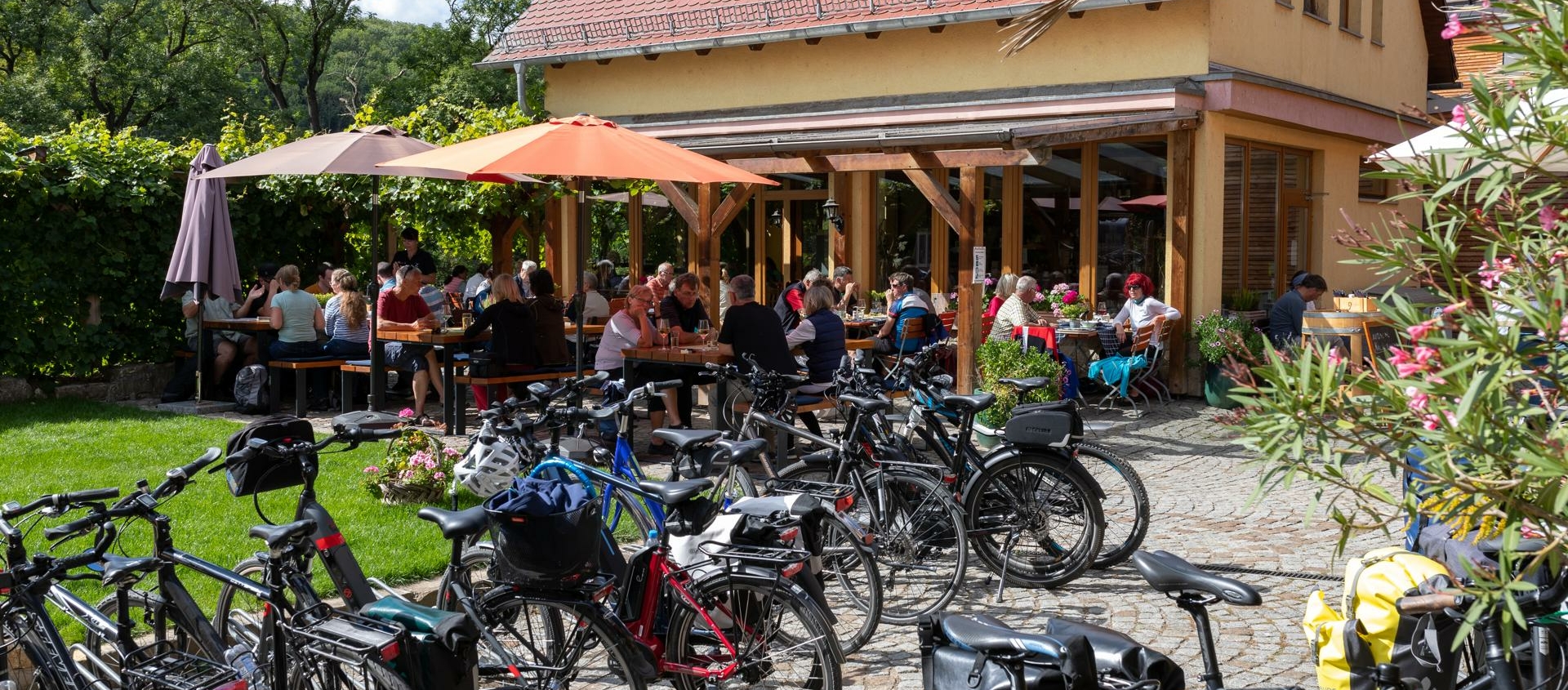 Mehrere Fahrräder stehen auf einem gepflasterten Platz vor einem gelben Gebäude mit rotem Dach, im Hintergrund sitzen viele Menschen an Tischen unter Sonnenschirmen im Freien