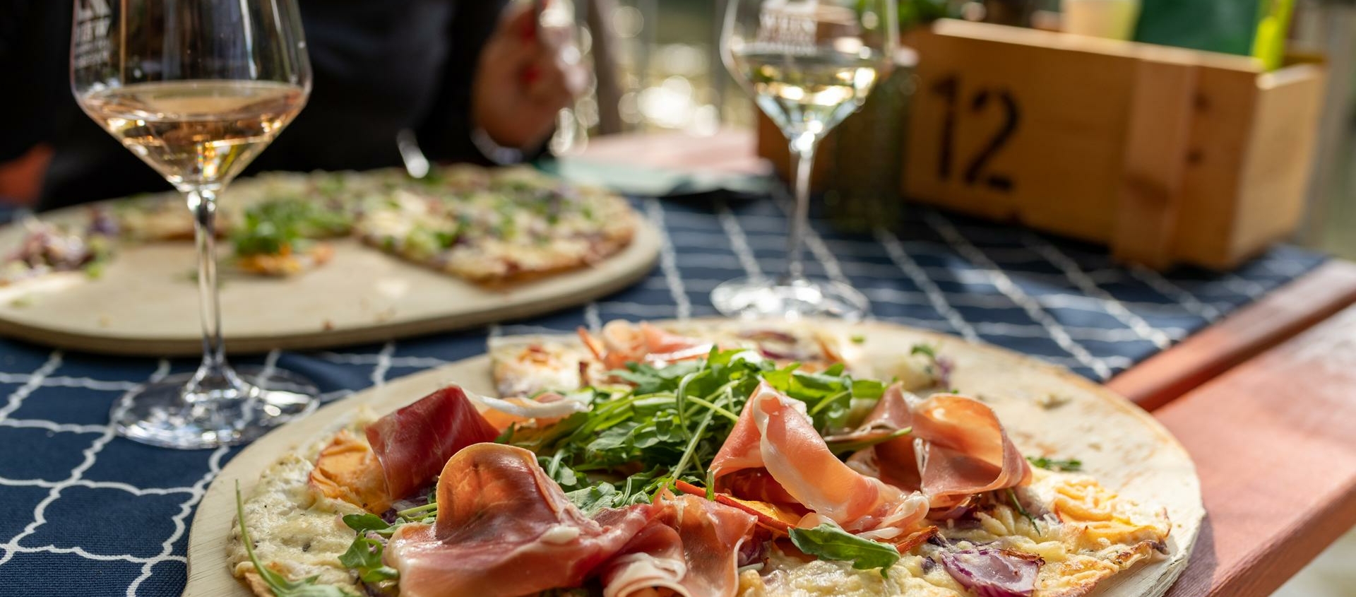 Flammkuchen mit Rucola und Schinken auf Holzbrett neben Weinglas auf blau-weiß kariertem Tischtuch im Freien