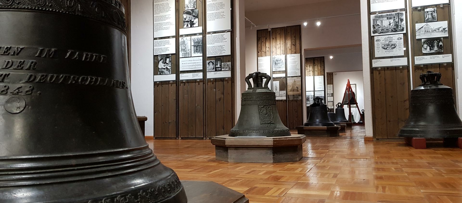 Mehrere historische Glocken unterschiedlicher Größe stehen auf Holzpodesten im Glockenmuseum in Apolda. An den Wänden erkennt man hohe Informationstafeln.