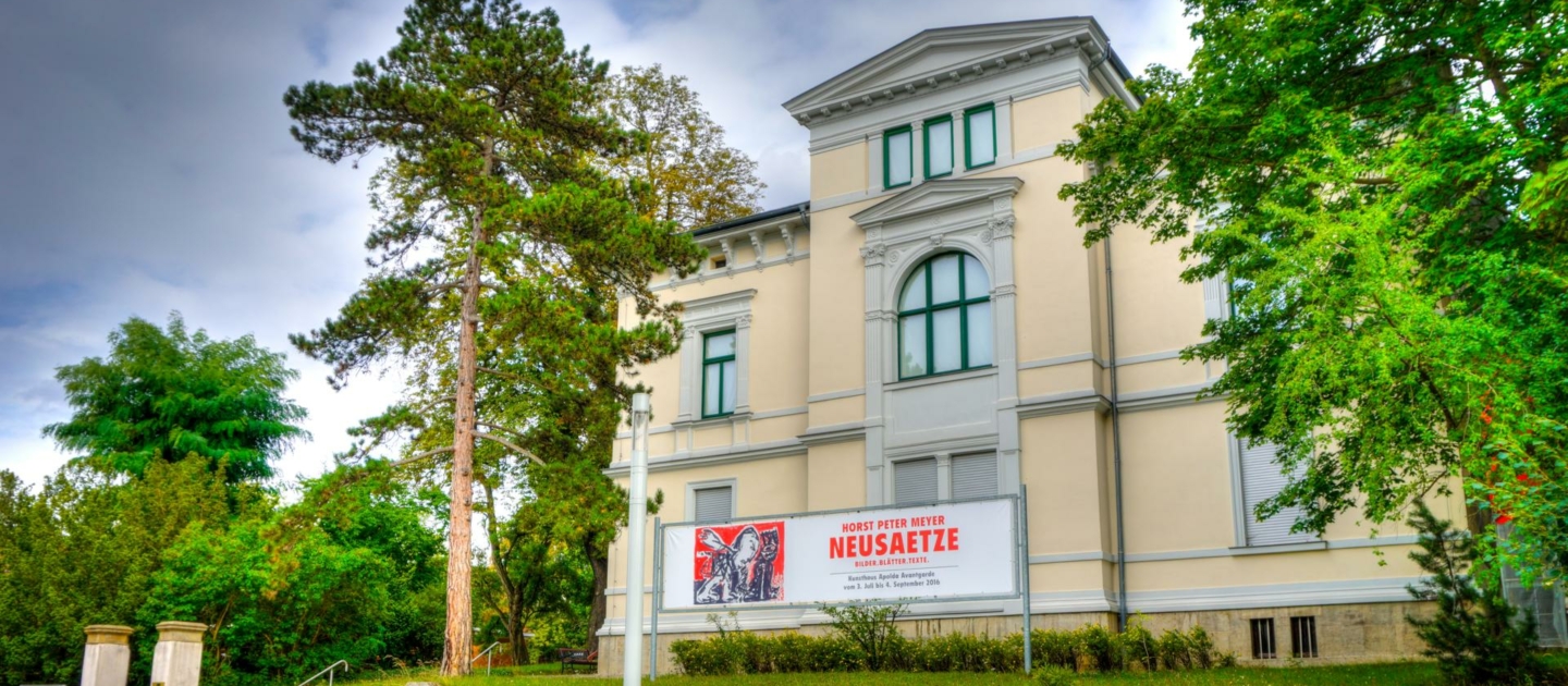 Das Kunsthaus Avantgarde Apolda, ein historisches Gebäude mit klassizistischer Fassade, umgeben von Bäumen und einem Steinmauerabsatz mit einem Banner für die Ausstellung 'NEUSÄTZE'