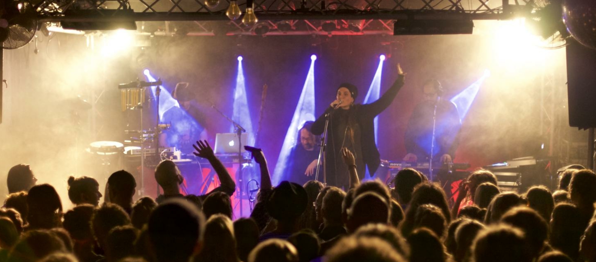 Bühne im Kulturbahnhof Jena mit Sängerin und Band vor Publikum, beleuchtet von bunten Scheinwerfern und Nebel.