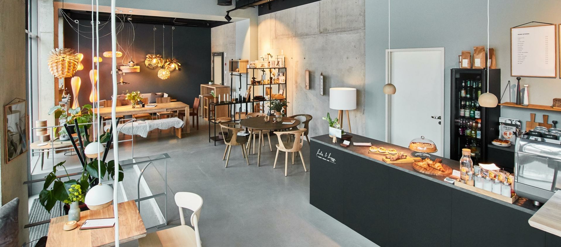 Moderner Caféinnenraum mit Holzstühlen, Tischen, schwarzer Theke mit Gebäck, Regalen mit Produkten und hängenden Lampen in einem Raum mit Sichtbetonwänden und sichtbaren Lüftungsrohren