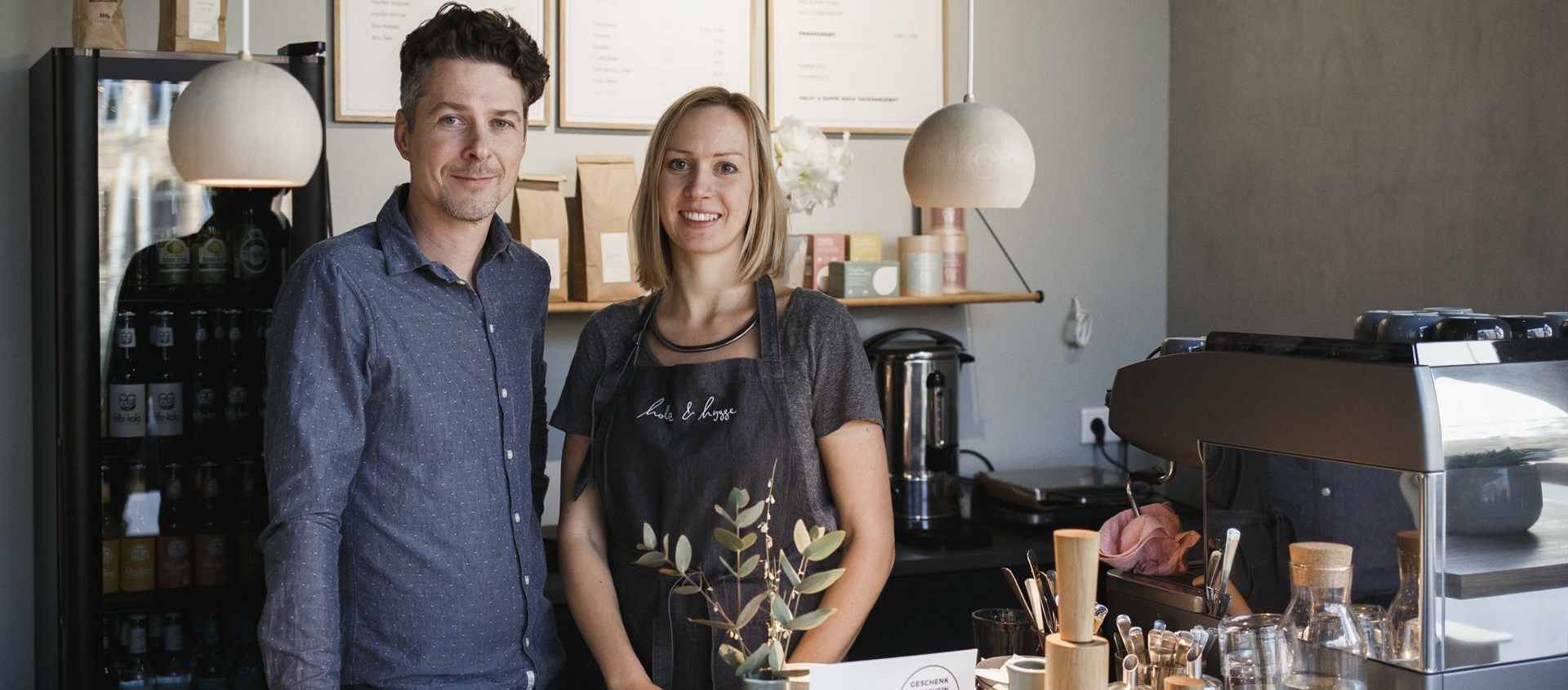 Zwei Personen stehen hinter einer Theke mit Kaffeebar-Utensilien und Pflanzen in einem Café mit drei gerahmten Menüs an der Wand.
