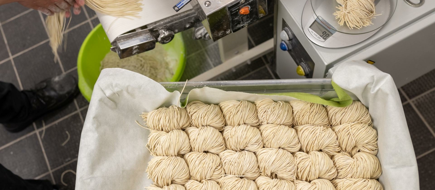 Mehrere kleine Portionen frischer Ramen-Nudeln liegen auf einem mit Tuch ausgelegten Tablett, daneben hält eine Person eine Portion Nudeln neben einer Nudelmaschine und einer digitalen Waage.