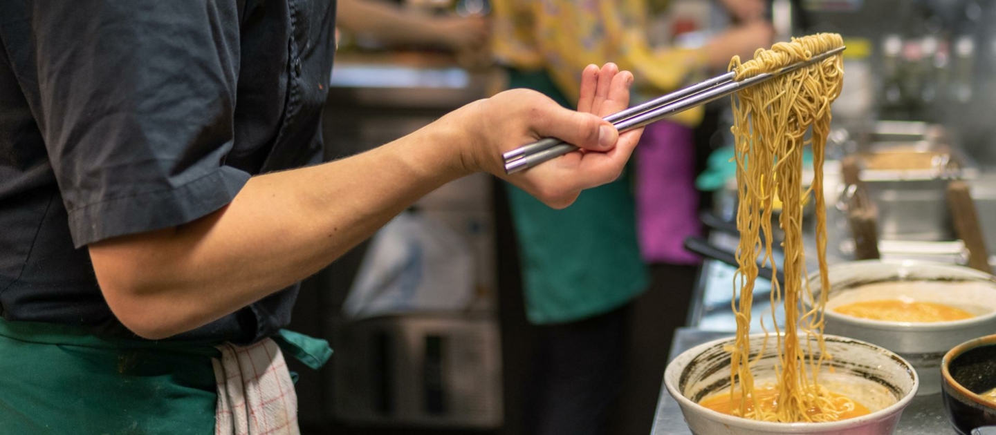 Koch in schwarzer Kochkleidung und schwarzer Mütze hebt mit Essstäbchen Ramen-Nudeln aus einer Schüssel in einer japanischen Ramen-Küche in Jena