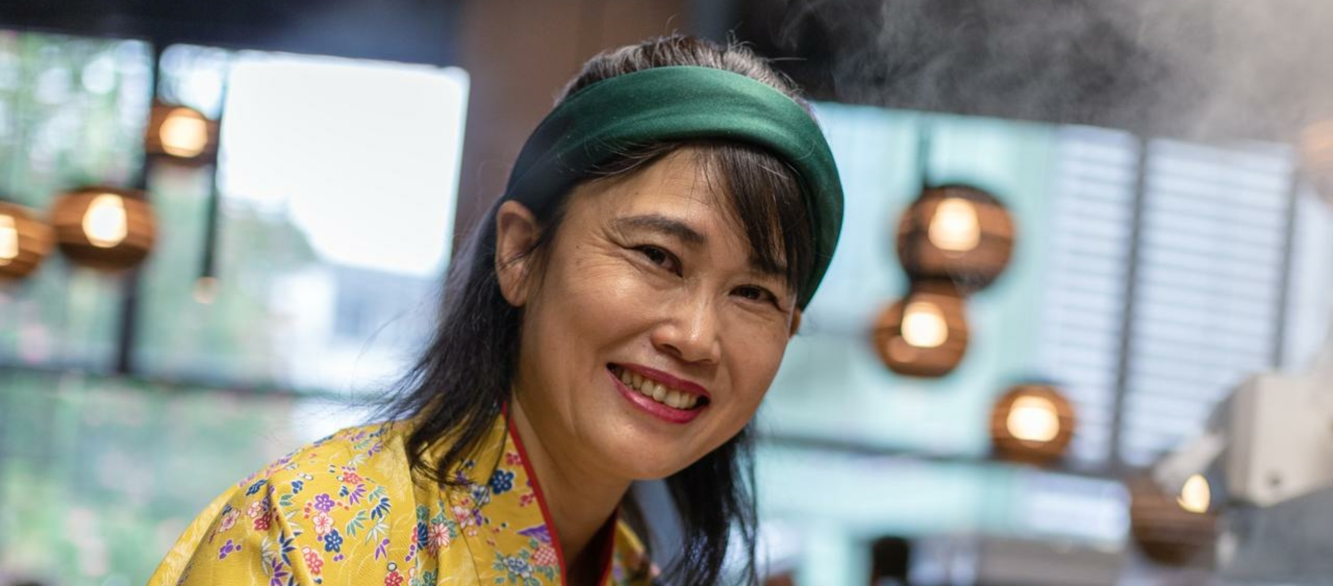 Person in gelbem, geblümtem Kimono und grünem Stirnband bereitet in der Küche des Jen Ramen in Jena eine Schüssel mit Ramen zu