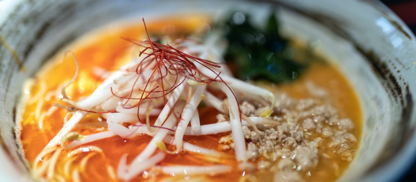 Nahaufnahme einer Schüssel japanischer Ramen mit dünnen Nudeln, Mungobohnensprossen, gehacktem Fleisch, grünen Algen und roten Chilifäden in einer Brühe