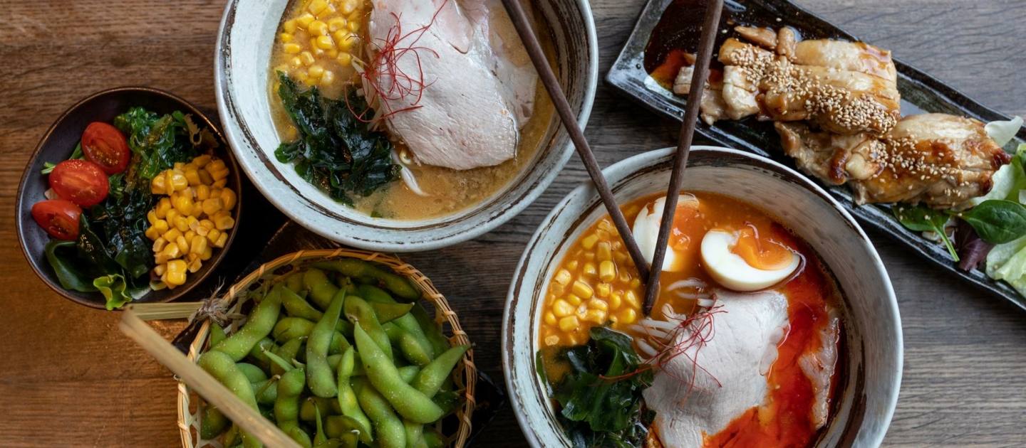 Zwei Schalen japanischer Ramen mit Brühe, Schweinefleisch, Mais, Algen und Chilifäden, daneben ein Korb mit Edamame, ein Teller mit gegrilltem Fleisch und eine kleine Schale mit Tomaten, Mais und Algen auf Holztisch im Jen Ramen in Jena.