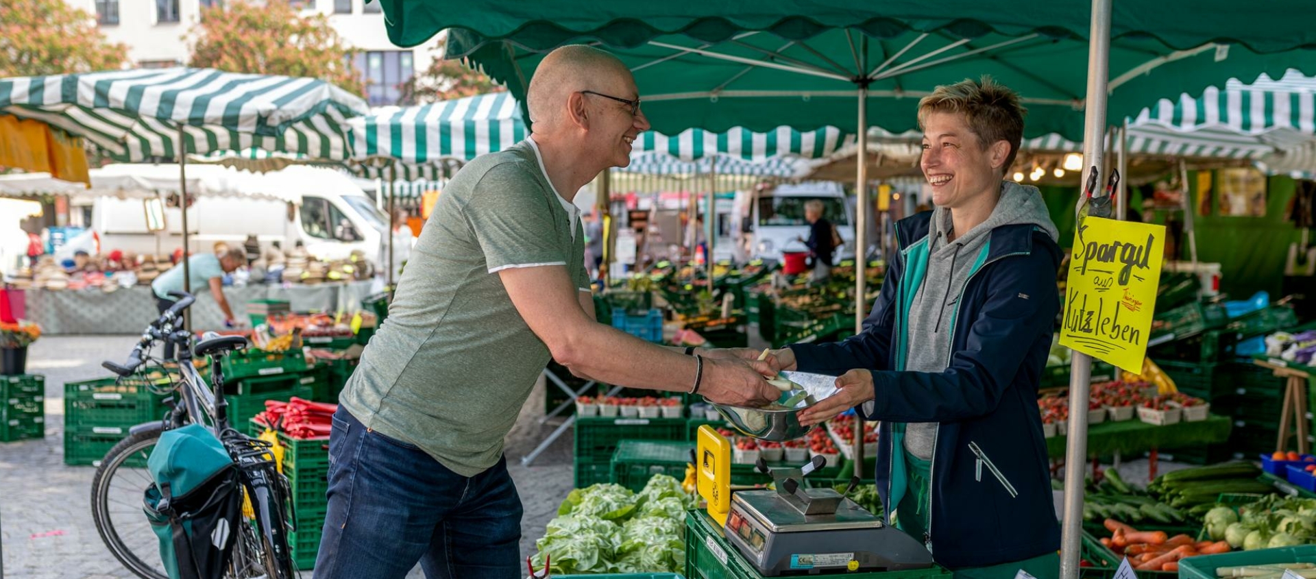 Zwei Personen tauschen an einem Marktstand mit grün-weiß gestreiftem Sonnenschutz Gemüse aus, im Hintergrund weitere Marktstände und ein Fahrrad.