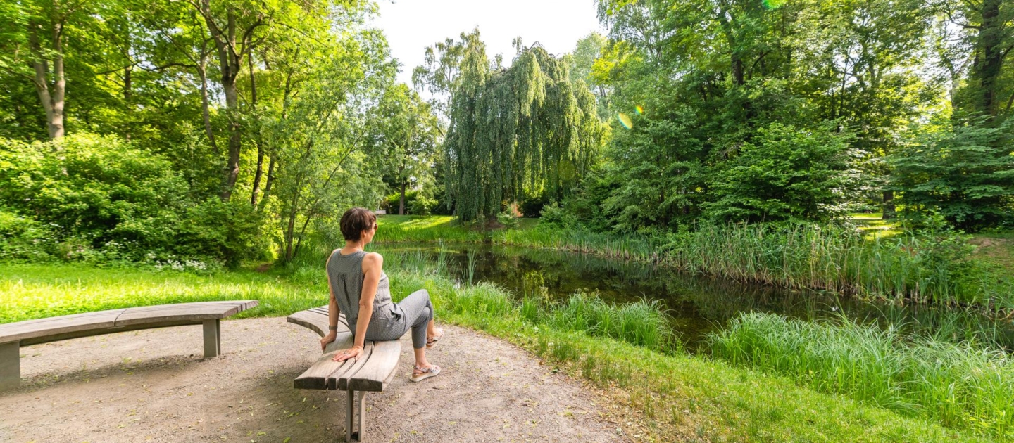 Frau sitzt auf einer Parkbank im Paradiespark in Jena und blickt auf einen Teich mit umgebender grüner Vegetation