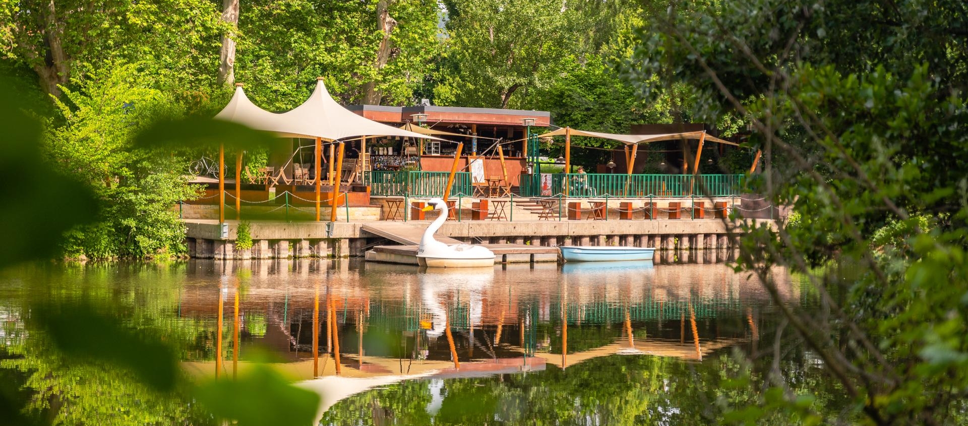 Holzsteg mit Sitzgelegenheiten und Sonnensegeln am Ufer der Saale, davor ein weißes Tretboot in Schwanform, umgeben von dichtem Grün in Jena