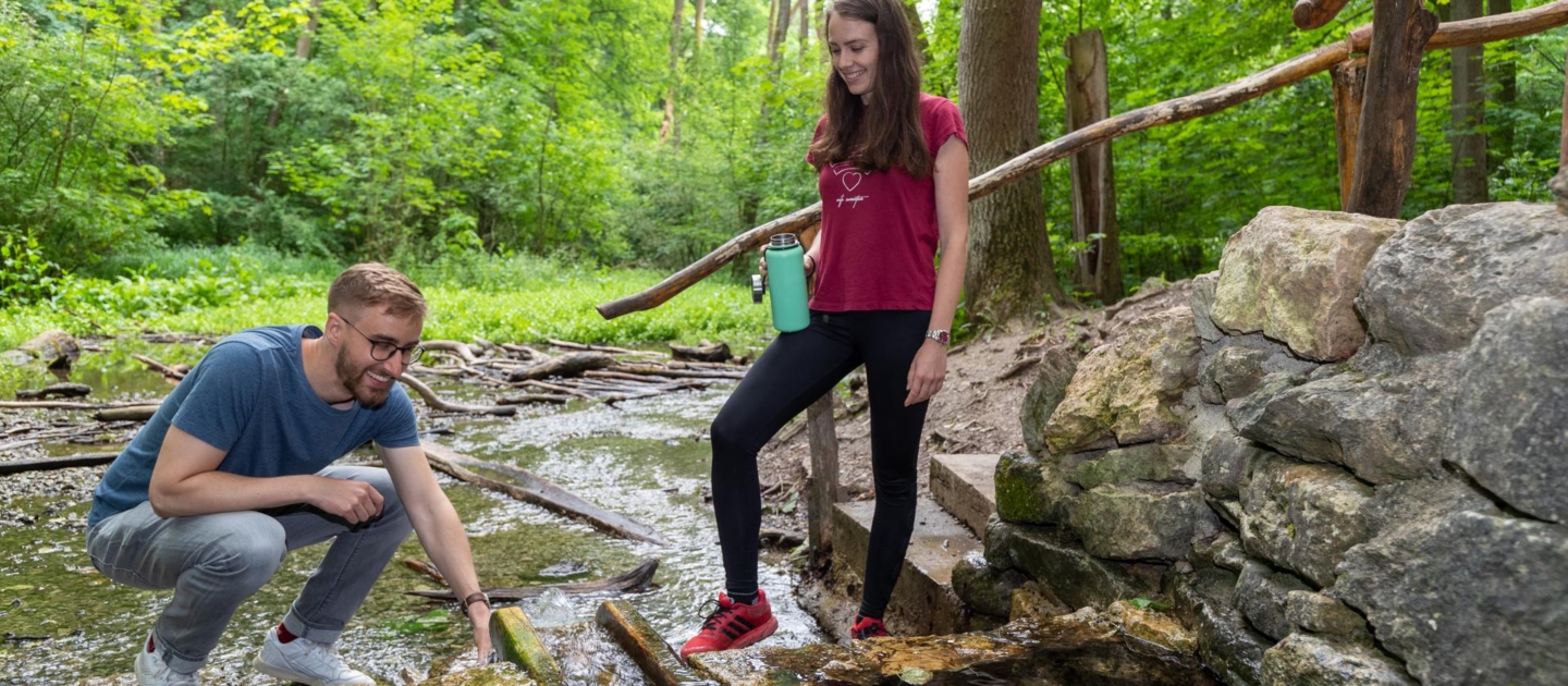 Zwei junge Personen an einem Bachlauf am Fürstenbrunnen in Jena, eine hockt und berührt das Wasser, die andere steht mit Trinkflasche auf einem Stein neben einer Steinmauer