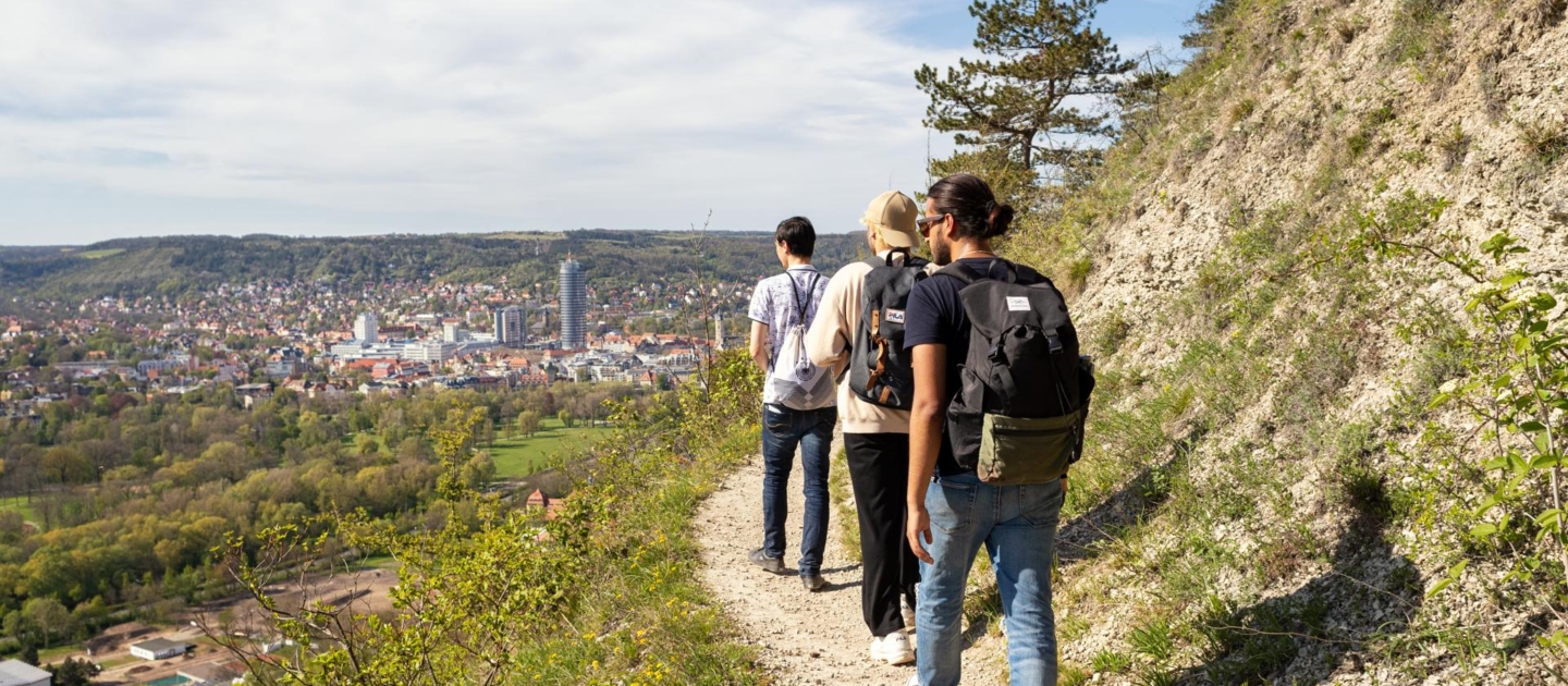 Vier Wanderer mit Rucksäcken gehen auf einem schmalen Pfad der SaaleHorizontale mit Blick auf die Stadt Jena