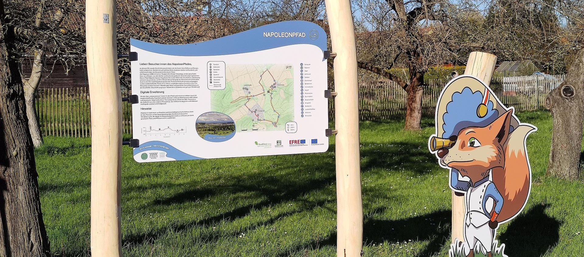 Informationsschild zum Napoleonpfad in Jena zwischen zwei naturbelassenen Holzpfosten mit einer Karte und Text sowie eine stehende Figur eines Fuchses in historischer Uniform auf einer Wiese mit Bäumen im Hintergrund