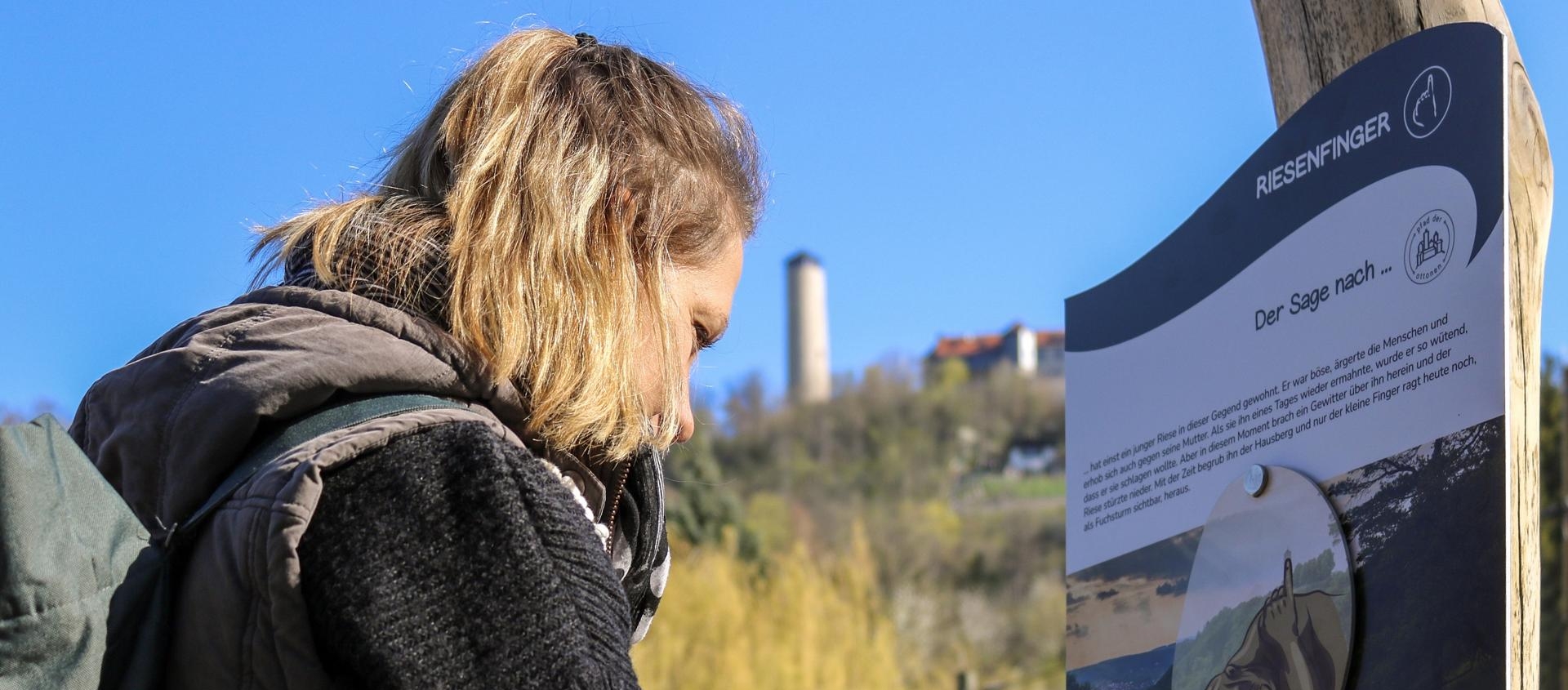 Person mit Rucksack liest Informationstafel zum Pfad der Ottonen in Jena im Freien mit Industriegebäude im Hintergrund