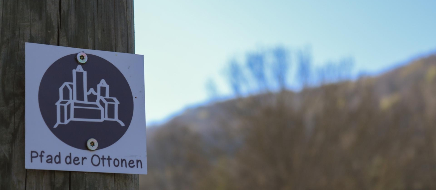 Holzpfahl mit einem quadratischen Schild, das eine stilisierte Burg und den Schriftzug 'Pfad der Ottonen' zeigt, unscharfer Hintergrund mit Bäumen und Hügeln.