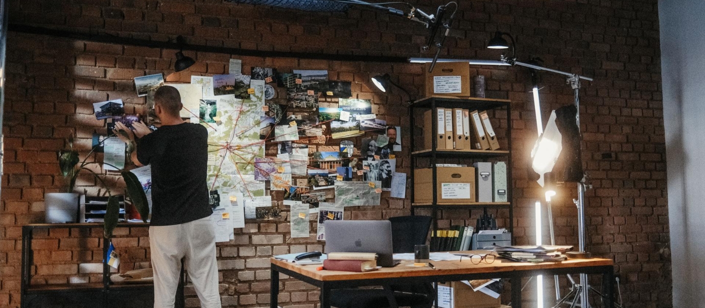 Person steht vor einer Pinnwand mit Fotos und Karten, verbindet Punkte mit roten Fäden in einem Büro mit Backsteinwand