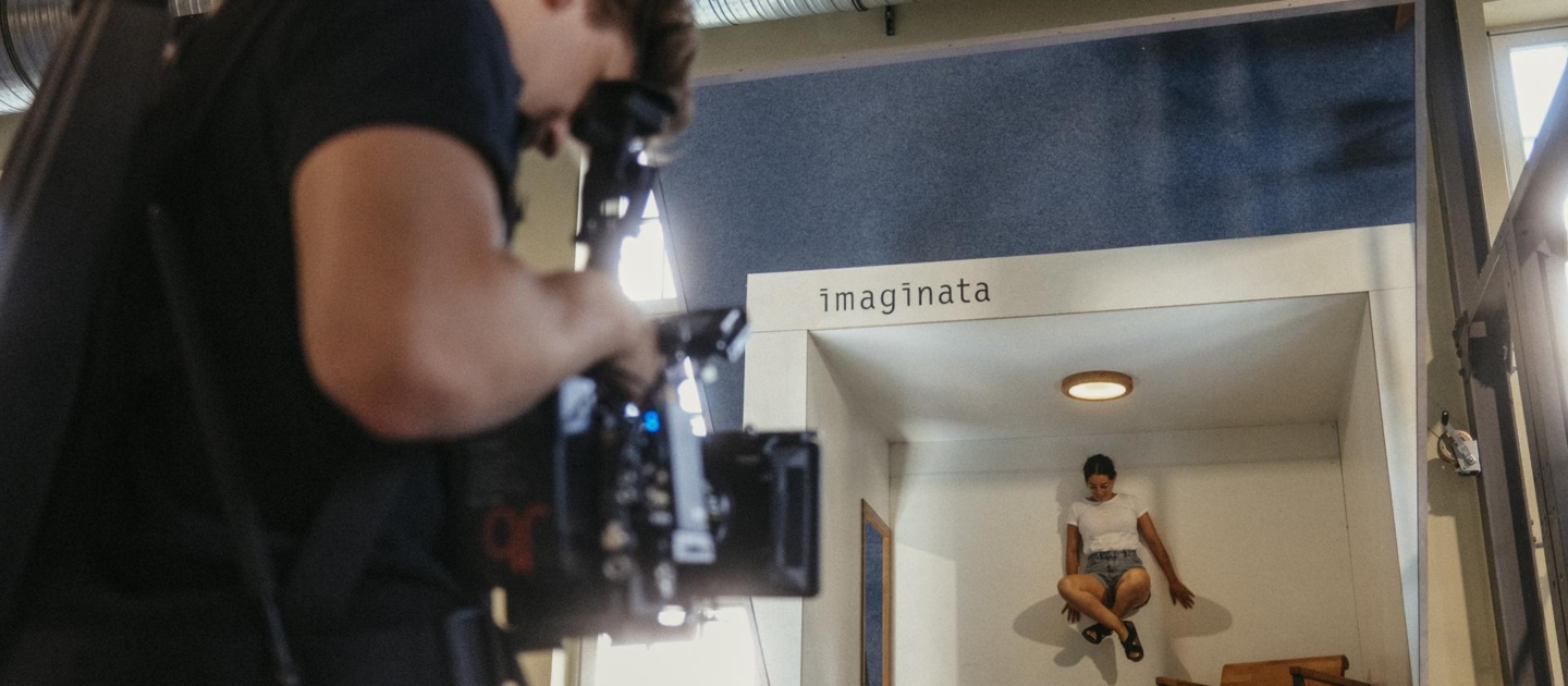 Mann filmt Frau, die in einem kleinen weißen Raum mit dem Schriftzug 'imaginata' an der Wand scheinbar schwebt