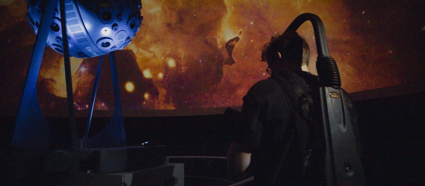 Person mit Staubsauger-Rucksack vor einem Planetariumsprojektor und einer Projektion eines farbigen Weltraumnebels in Jena