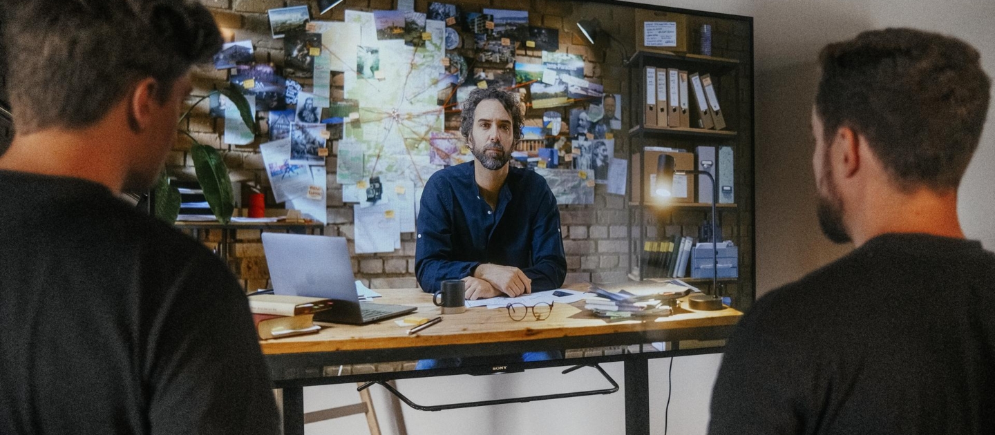 Zwei Männer sitzen vor einem Tisch und hören einem dritten Mann zu, der an einem Tisch mit Papieren und Laptop vor einer Wand mit Notizen und Fotos sitzt