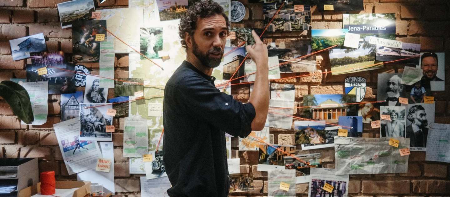 Person steht vor einer Wand mit zahlreichen Fotos, Notizen und roten Fäden, die Verbindungen zwischen den Elementen zeigen