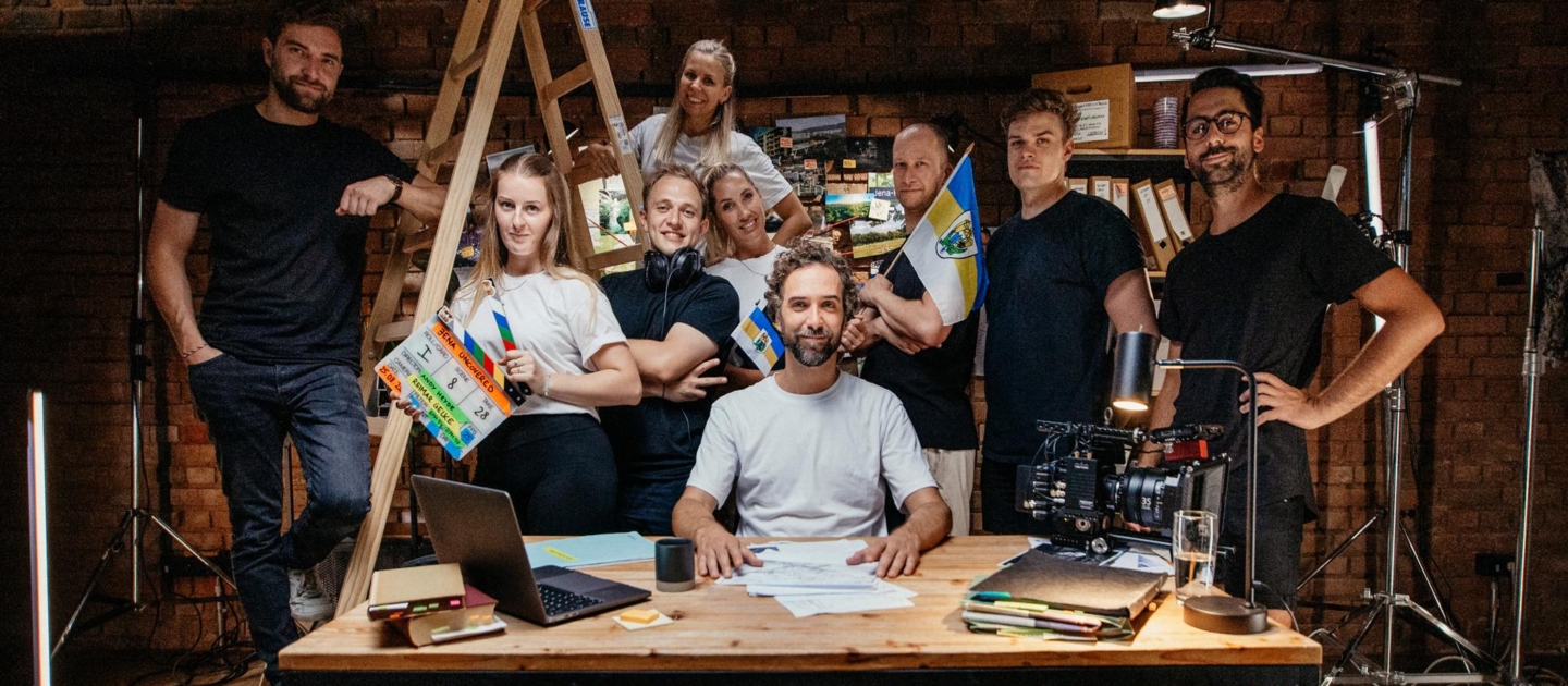 Acht Personen in einem Studio mit Film- und Beleuchtungsausrüstung, eine Person sitzt an einem Holztisch mit Laptop und Papieren, andere stehen um sie herum