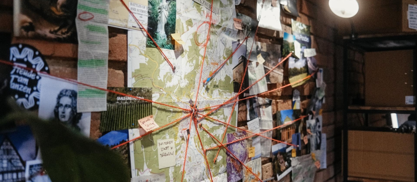 Wand mit Landkarte und zahlreichen Fotos, Notizen und roten Fäden, die verschiedene Punkte auf der Karte verbinden