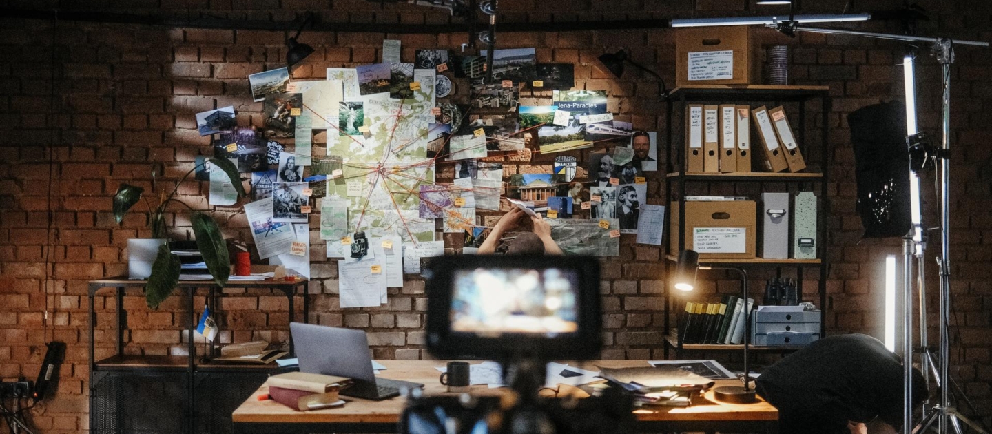 Arbeitszimmer mit Pinnwand voller Karten und Fotos, Person sitzt am Tisch, Kamera im Vordergrund, Regale mit Ordnern und Beleuchtung