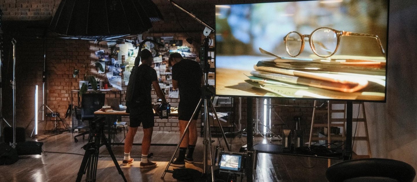 Zwei Personen in einem Studio mit Kameraausrüstung vor einer großen Leinwand, die eine Nahaufnahme von Brille auf einem Stapel Bücher zeigt