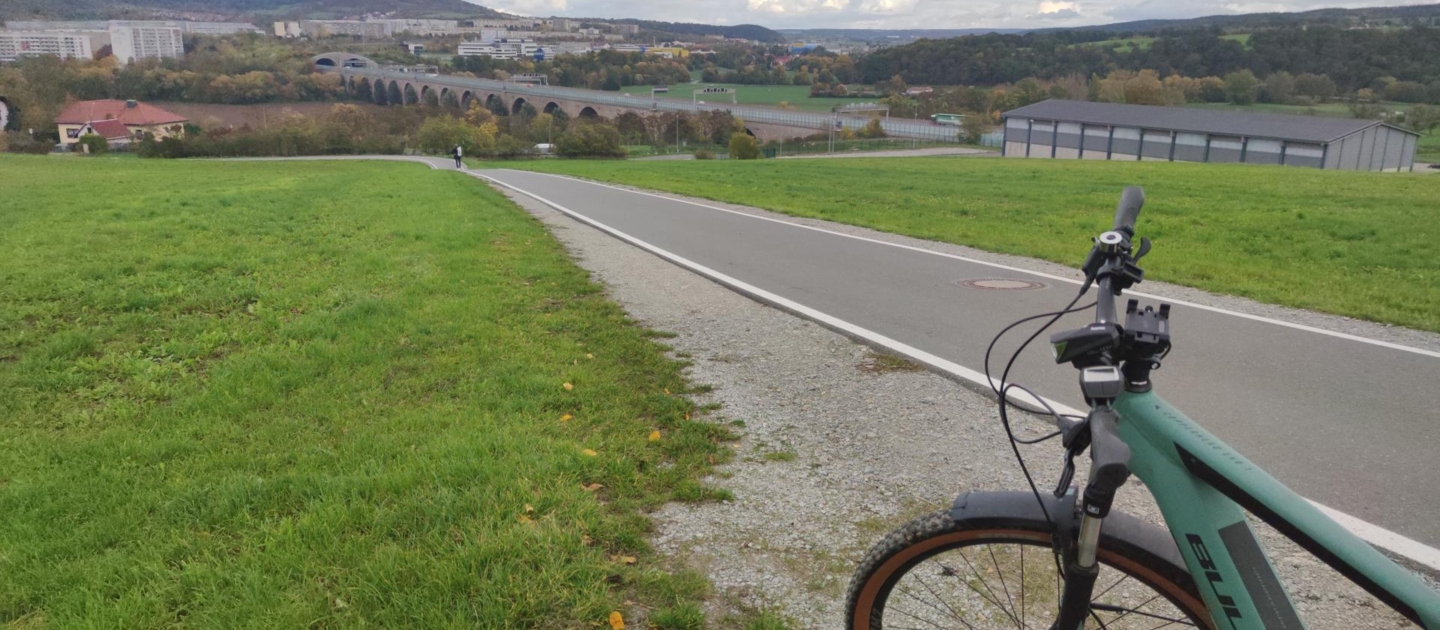 Blick auf eine asphaltierte Radstrecke mit grünem Mountainbike im Vordergrund, im Hintergrund Jena mit Viadukt und bewaldeten Hügeln unter bewölktem Himmel