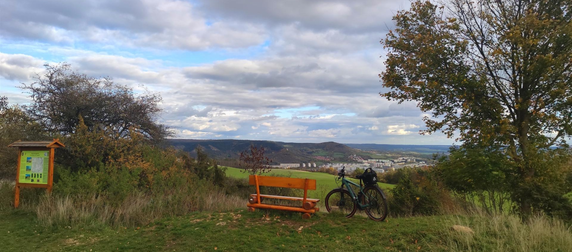 Fahrrad neben einer Holzbank auf einem Hügel mit Blick auf Jena und bewölktem Himmel