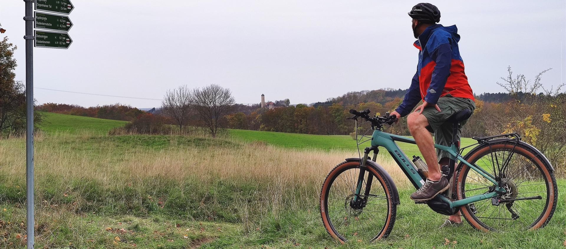 Mann auf Mountainbike blickt auf Wegweiser in einer grünen Landschaft bei Jena, im Hintergrund der Fuchsturm