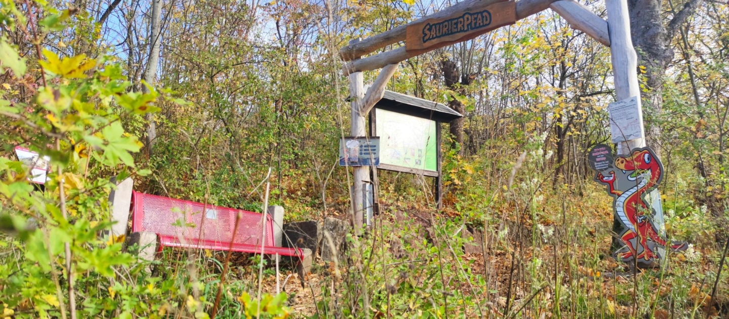 Eingang zum Saurierpfad in Jena mit hölzernem Tor, roter Bank und Infotafel im herbstlichen Wald