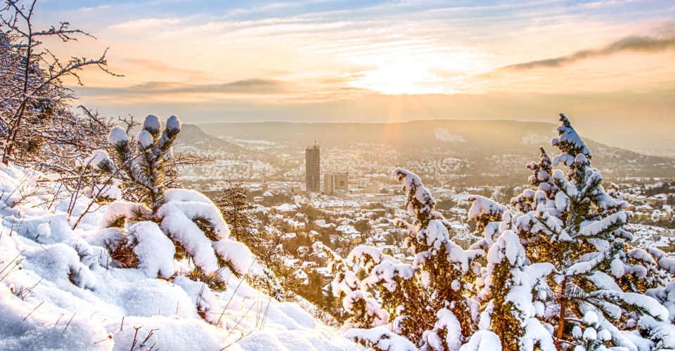 Winterliche Stadtansicht von Jena mit schneebedeckten Bäumen im Vordergrund und Sonnenuntergang über der Stadt