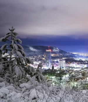 Blick auf die verschneite Stadt Jena bei Nacht mit beleuchteten Gebäuden, schneebedeckten Bäumen im Vordergrund und rauchenden Schornsteinen im Hintergrund