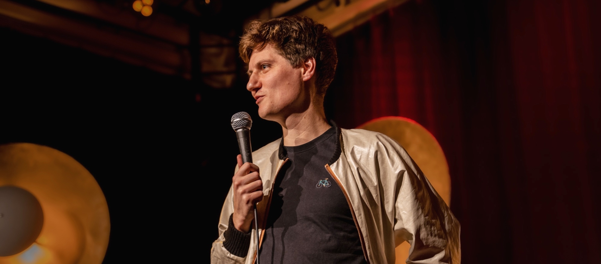 Friedrich Herrmann beim Poetry Slam, spricht in ein Mikrofon, trägt helle Jacke und schwarzes T-Shirt mit Fahrradmotiv, Bühnenbeleuchtung im Hintergrund