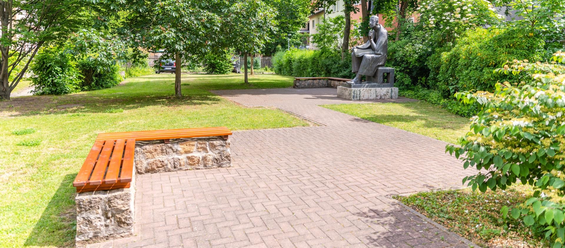 Skulptur von Albertus Magnus sitzend mit Buch im Frommannschen Anwesen in Jena, umgeben von Bäumen und Sitzbänken