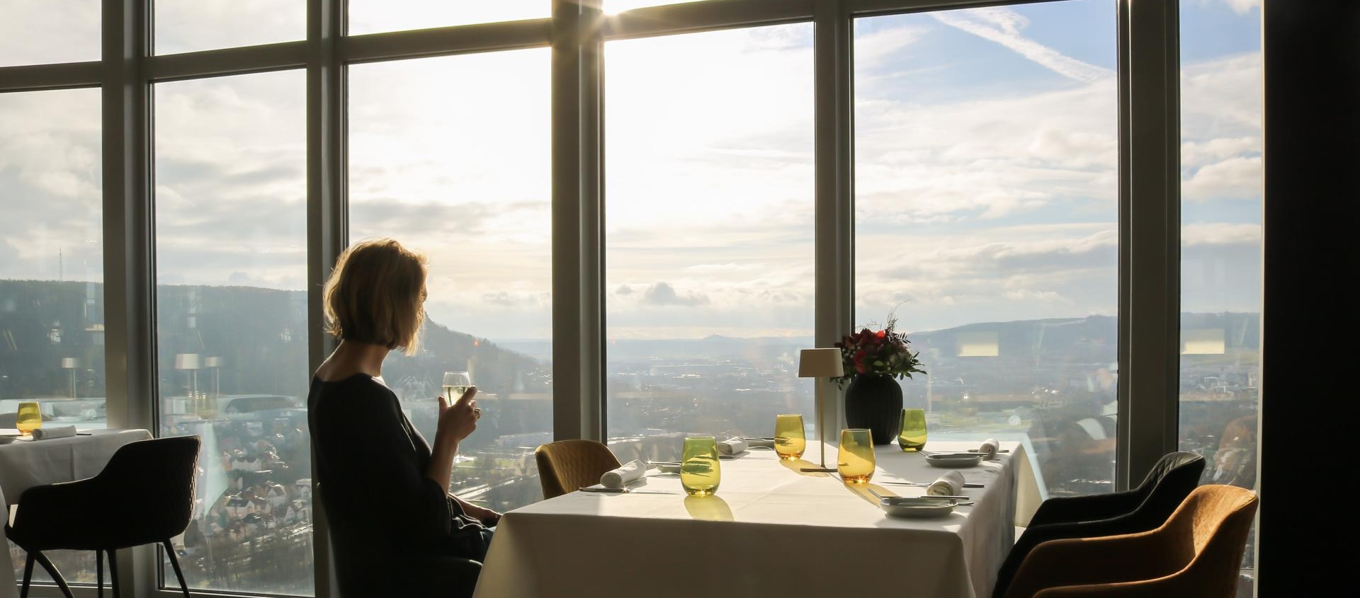 Frau sitzt im Restaurant des Jentowers in Jena, trinkt aus einem Glas, großer Panoramablick auf Stadt und Hügel bei Sonnenuntergang