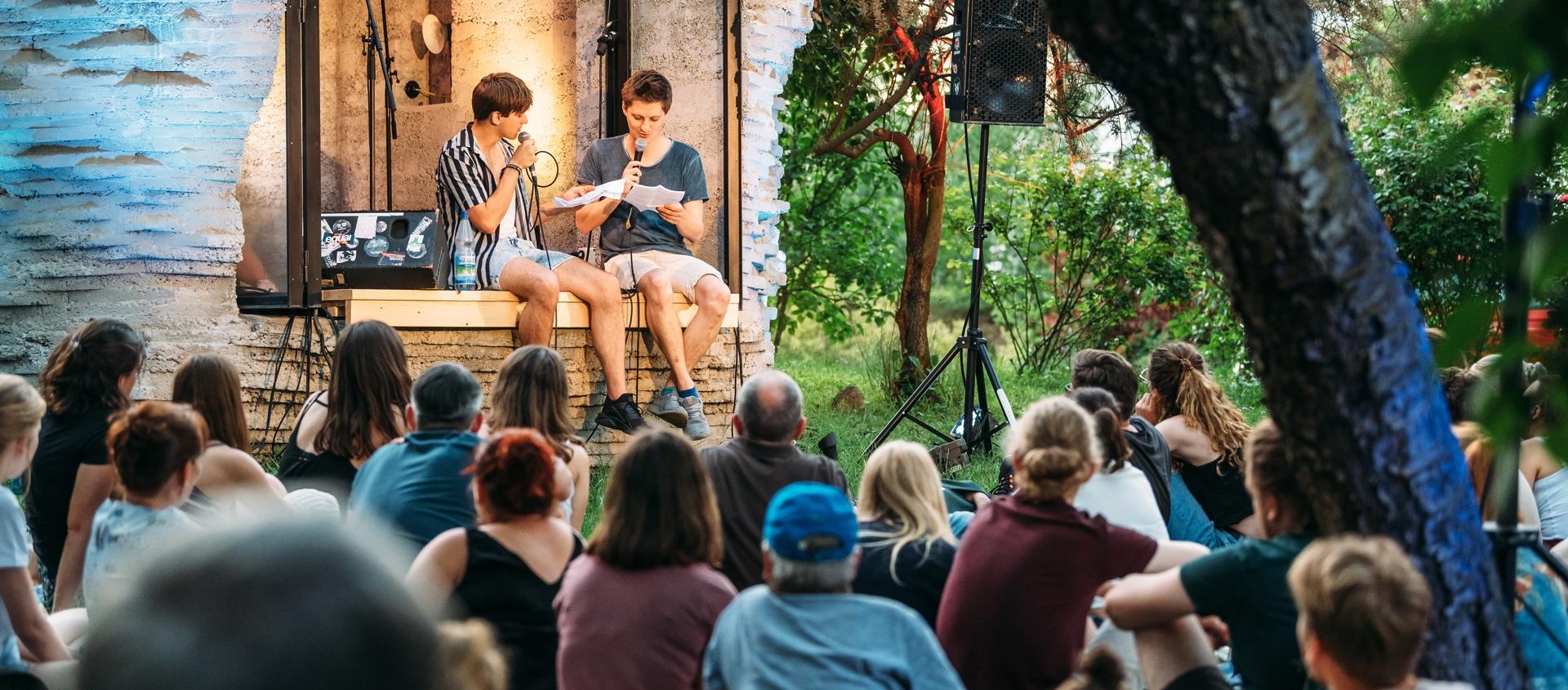 Poetry Slam mit Friedrich Herrmann im Garten der Villa Rosenthal, zwei junge Männer sitzen auf einer Bühne und lesen vor, Publikum sitzt auf dem Rasen