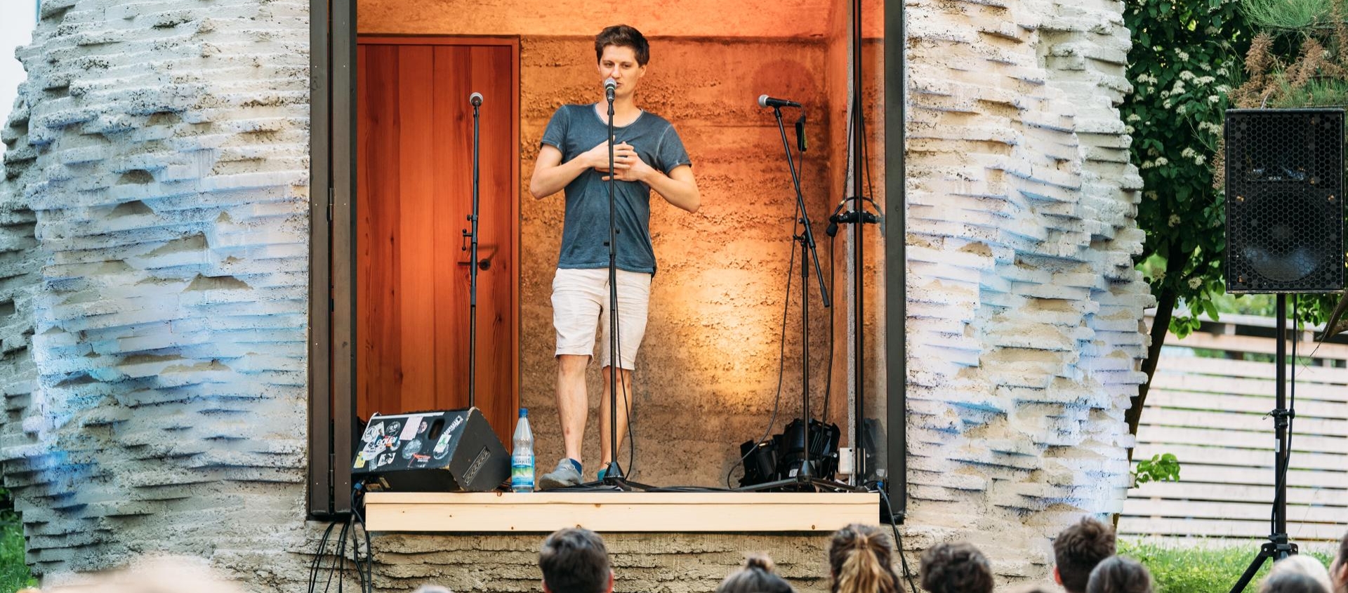 Ein junger Mann in Shorts und T-Shirt steht auf einer kleinen Bühne vor einem Publikum und spricht in ein Mikrofon bei einem Poetry Slam im Freien
