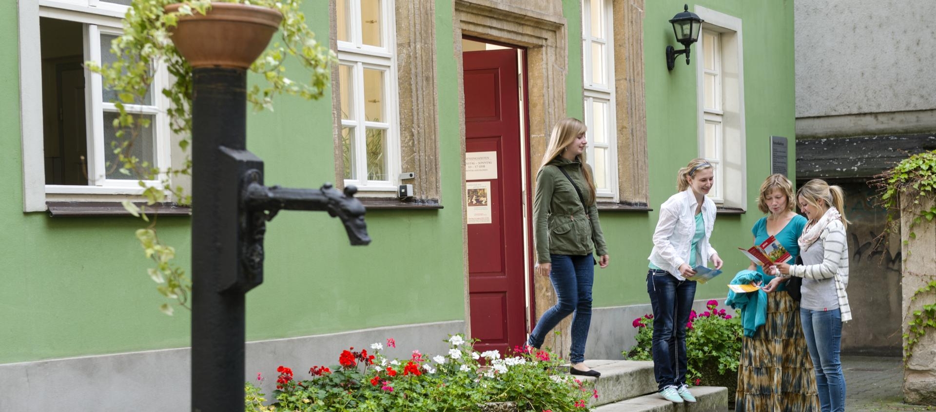 Vier Frauen stehen vor dem grünen Romantikerhaus in Jena neben einem alten Brunnen und betrachten gemeinsam eine Broschüre