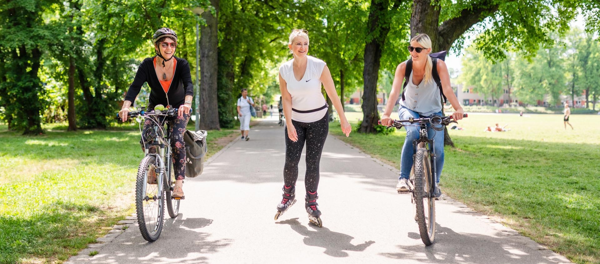 Drei Frauen im Volkspark Oberaue in Jena, zwei fahren Fahrrad, eine fährt Inliner auf einem von Bäumen gesäumten Weg