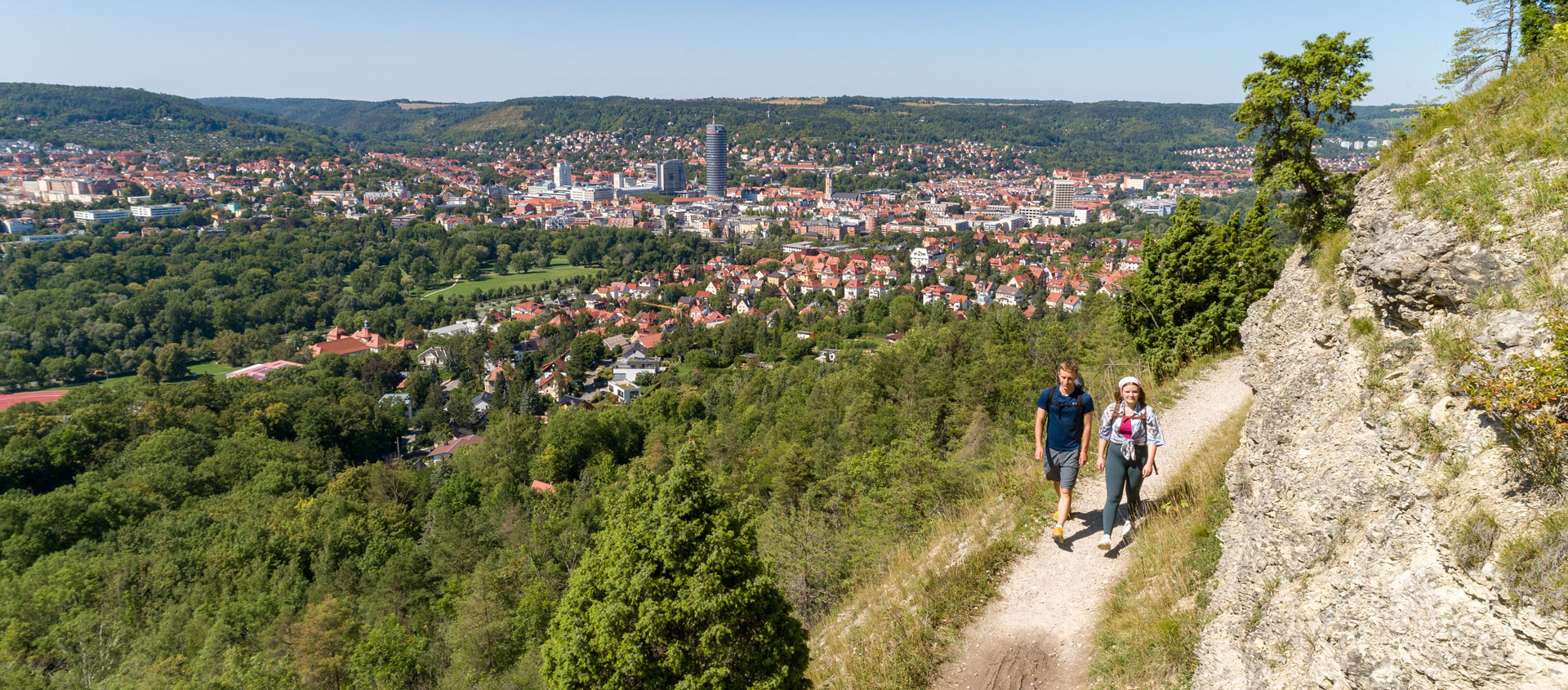Zwei Wanderer auf dem Saalehorizontale-Wanderweg mit Blick auf die Stadt Jena und bewaldete Hügel unter klarem Himmel