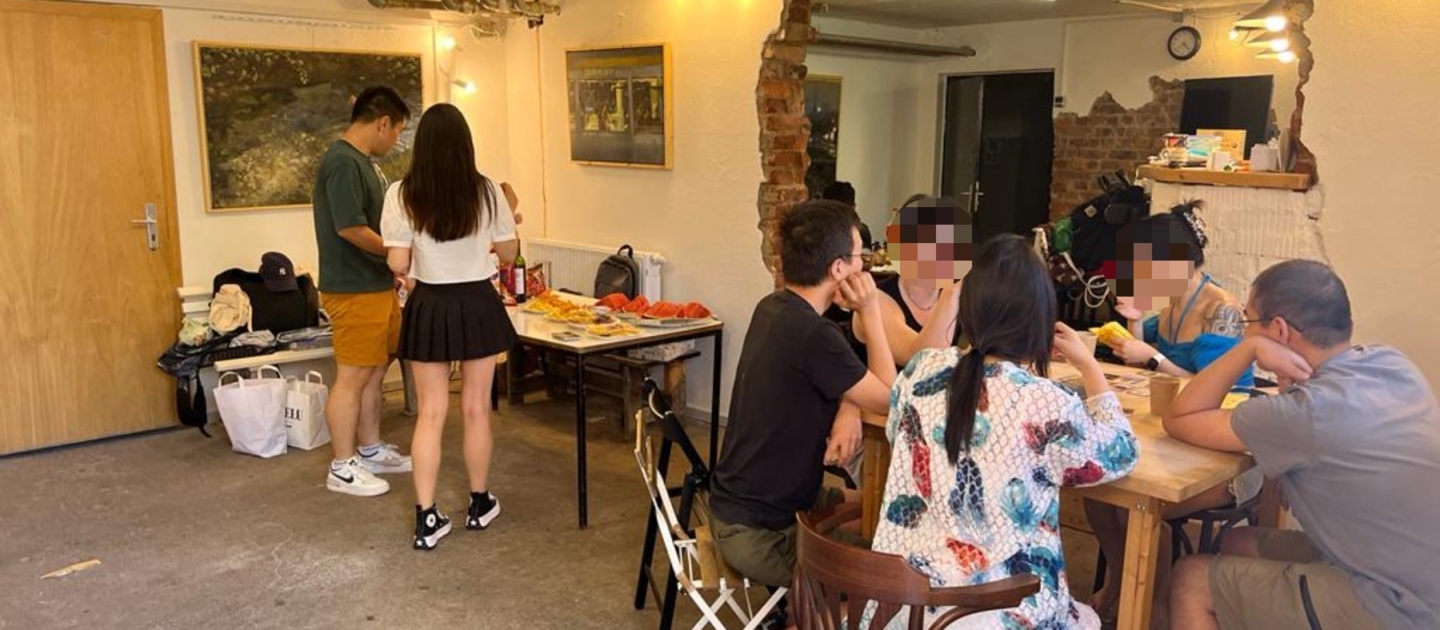 Menschen sitzen und stehen in einer Galerie in Jena, einige essen und unterhalten sich an einem Holztisch, andere stehen an einem Tisch mit Snacks und Getränken