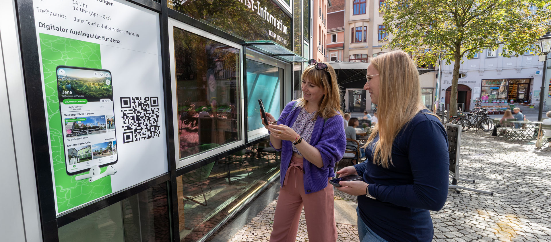 Zwei Frauen scannen vor der Jena Tourist-Information einen QR-Code auf einem Plakat für eine digitale Stadtführung.